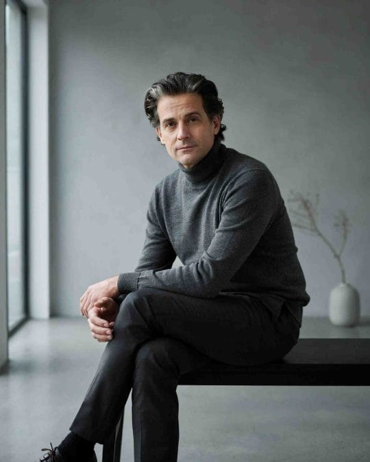 Mature man with brushed-back wavy hair wearing a charcoal sweater.