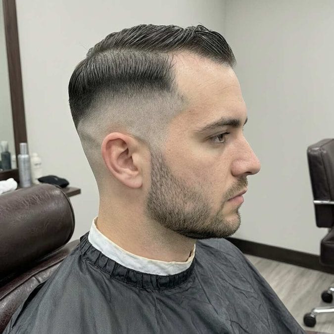 Man with a low taper fade, short side part, and neat beard in a barbershop setting.