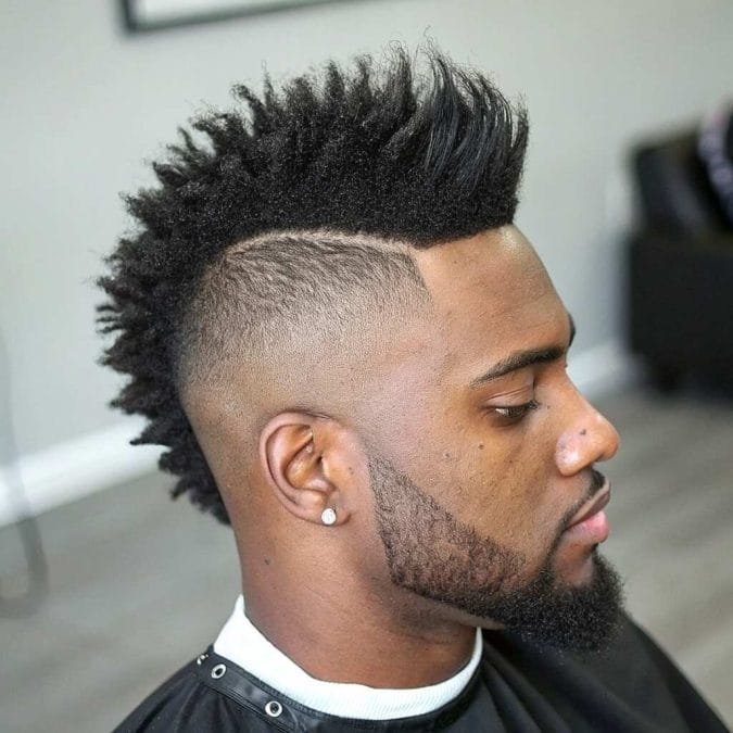 Burst fade mohawk haircut with textured natural hair, a sharp surgical part, and a defined beard for a bold modern look.