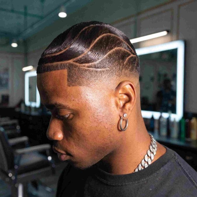 Drop fade haircut with styled waves and intricate shaved design details for a polished appearance.