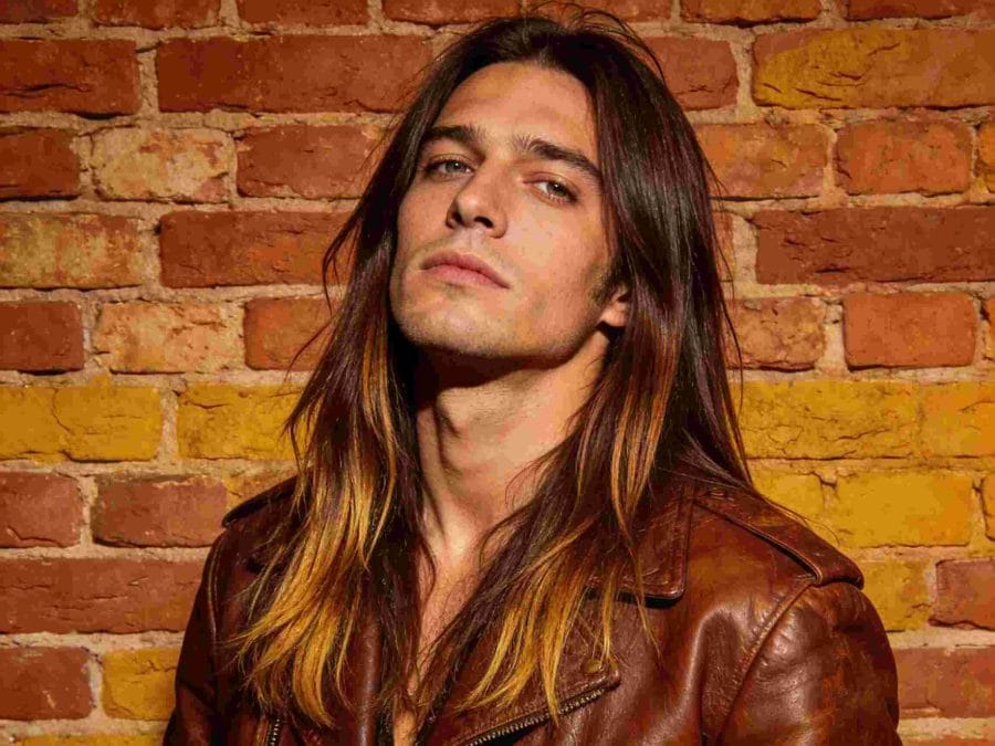 Man with long straight rocker hair, a middle part, and lighter ends against a brick wall.